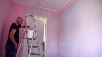 homem pinta as paredes cor-de-rosa na sala para a cor magenta com um rolo de pintura em uma escada video