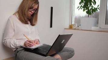 trabajo remoto. la mujer toma notas en el cuaderno e imprime en el teclado de la computadora portátil. educación a distancia y concepto de aprendizaje electrónico video
