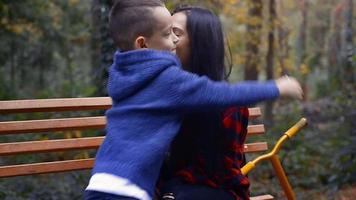 Young preschooler boy kiss his happy mother at her nose on a bench at the park at warm autumn day video