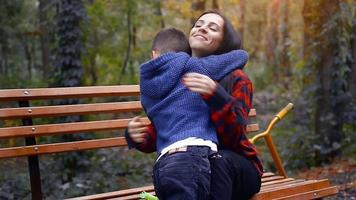 Young preschooler boy kiss his happy mother at her nose on a bench at the park at warm autumn day video