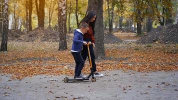 mom helps her son to ride on a scooter at the warm autumn day in the park video