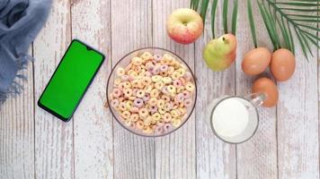 Top view of colorful cereal in a bowl, smart phone, milk, eggs and fresh fruits on the table video