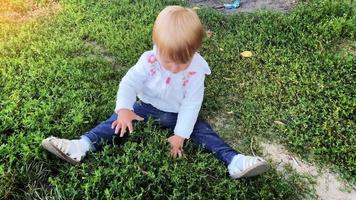 Charming little blonde caucasian toddler girl sits and cutting a grass on a lawn