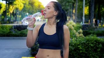jolie brune caucasienne sportive boit de l'eau après s'être entraînée à l'extérieur dans le parc lors d'une chaude journée d'été video