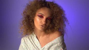 Beautiful young girl with fluffy curly hair and colourful makeup in studio on purple background in slow motion video