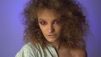 Beautiful young girl with fluffy curly hair and colourful makeup in studio on purple background in slow motion video