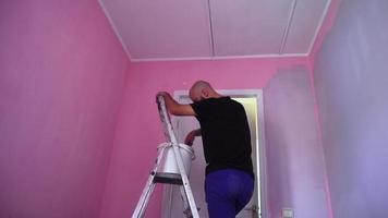 man paints the pink walls in the room to magenta color with a paint roller on a ladder video