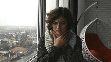 una mujer con un suéter y una bufanda sentada junto a la ventana con dolor de garganta en casa video