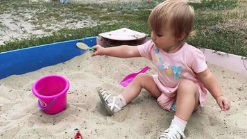 cute caucasian baby girl in pink dress plays in sandbox with shovel and bucket outdoors at summer time in 4k video