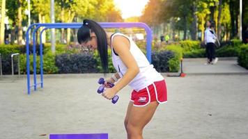 caucasian woman train her biceps muscles with dumbbells on a purple karemat at warm sunny day at the park video