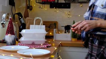 la mujer lo pone en la mesa y toma los contenedores del servicio de entrega de alimentos en la mesa cocina festiva escandinava blanca con decoración navideña. víspera de año nuevo, ahorrando tiempo, demasiado perezoso para cocinar, pedido caliente, caja de plástico