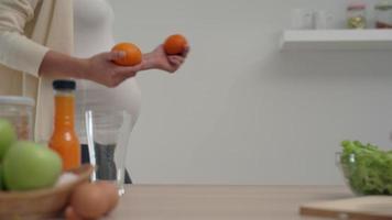 uma mulher grávida de bom humor joga alegremente laranja na sala da cozinha. mulheres grávidas se preparam para fazer suco de laranja para nutrir o feto. video
