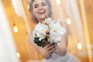 Beautiful bride with bouquet of flowers is looking through the window glass photo