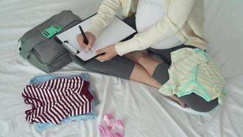 una mujer embarazada escribe en papel para volver a verificar necesario para el recién nacido. la madre está doblando y guardando la ropa para prepararse para el próximo bebé. feliz por el concepto de recién nacido. video