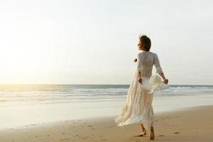 Woman wearing beautiful white dress is walking on the beach during sunset photo