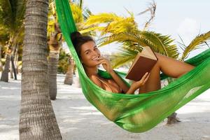 Happy and beautiful woman lying in the hammock and reading book photo