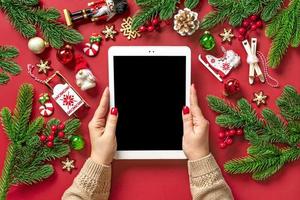 top view female hands holding tablet with black digital screen, Christmas box and decor on red table Flat lay Holiday shopping list, Happy New Year, online shop, chooses gifts, makes purchases Mockup photo