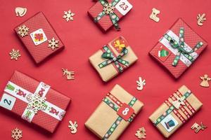 cajas de regalo rojas y verdes envueltas a mano decoradas con cintas, copos de nieve y números, adornos navideños y decoración en la mesa roja concepto de calendario de adviento de navidad vista superior tarjeta de vacaciones plana foto