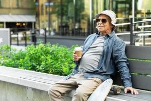 anciano con un longboard sentado en el banco y tomando café en un parque de la ciudad foto