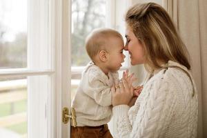 Mother and her little baby son wearing warm sweaters standing by the window photo