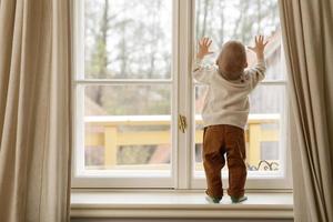 Baby boy is standing on the windowsill and looking at the window photo