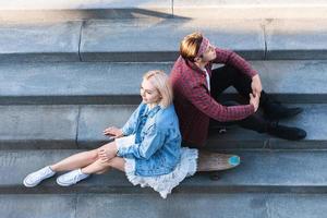 una elegante pareja adolescente con un longboard está sentada en unas escaleras de hormigón foto