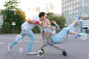 la pareja se divierte con un carrito de compras y hace burbujas en el estacionamiento de un supermercado foto