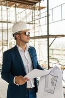 Arquitecto hombre vestido con traje formal y sombrero duro durante el control de la construcción de edificios sosteniendo planos en un sitio de construcción