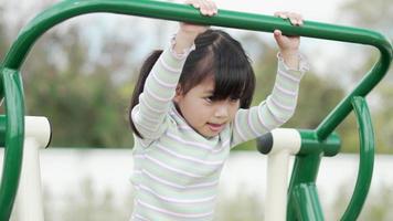 niña asiática disfruta jugando en equipos de ejercicio al aire libre con una sonrisa, en el patio de recreo