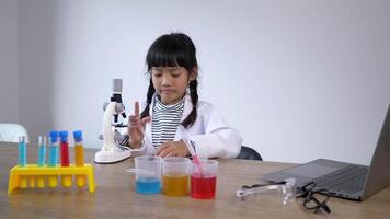 grappig meisje gebruik microscoop, laptop computer en apparaat van experimenteren met vloeistoffen Aan tafel, terwijl aan het studeren wetenschap chemie, selectief focus video
