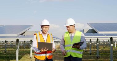 asiatischer Inspektor-Ingenieurmann, der ein Kontrollbrett hält, und junger Fachmann verwenden Laptop-Computer, zwei Ingenieure diskutieren während der Arbeit im Solarpark, Solarpanel-Station im Hintergrund video