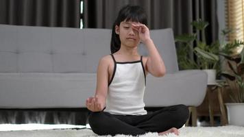 niña asiática sentada en la alfombra en pose de meditación en casa, cerró el ojo y puso la mano en la rodilla