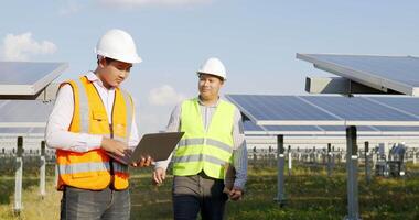 Asian Inspector Engineer man Holding checking board and young specialist man use laptop computer, Two engineers are discussing during working at solar farm, solar panel station in background video