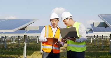 Asian Inspector Engineer man Holding checking board and young specialist man use laptop computer, Two engineers are discussing during working at solar farm, solar panel station in background video