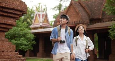 feliz pareja de viajeros asiáticos con la mano del sombrero juntos mientras visitan el antiguo templo. sonriente joven y mujer caminando y mirando templo antiguo. concepto de vacaciones, viajes y pasatiempos. video