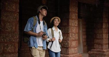 Happy asian traveler couple with hat takes a photo and visits ancient temple. Smiling young man and woman walking and looking ancient temple. Holiday, travel and hobby concept. video
