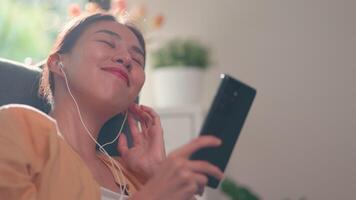 mujer asiática muy joven sentada y disfrutando escuchando música y cantando desde auriculares inalámbricos con teléfono inteligente en la sala de estar en casa, relajante y concepto de estilo de vida. video