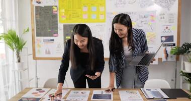 Two asian women checking to task on workplace desk while standing in office. One woman holding laptop and writing on paper, One woman using smartphone and writing notes on colorful sticky papers. video