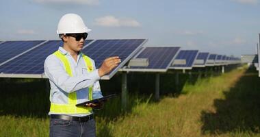 Handheld shot, Asian Young engineer man wearing white protection helmet and sunglasses standing at front of solar panel use digital pen write to record data on tablet while working in solar farm video