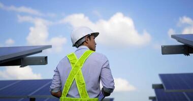 Handheld medium shot, rear view of Asian young inspector engineer man wearing helmet and hold report paper in hand walk to checking solar panel in solar farm video