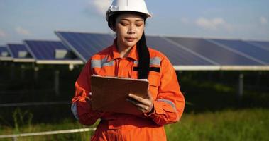 Handheld medium shot, Asian Young engineer woman wearing protection uniform and white helmet standing at front of solar panel to checking operation in paperwork while working in solar farm video
