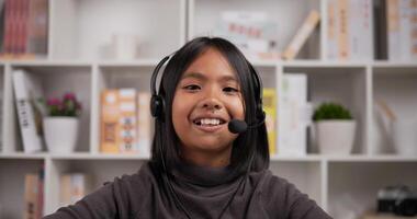 retrato de tiro na cabeça de fone de ouvido de menina sorridente asiática falando e olhando para a câmera enquanto está sentado na mesa em casa. jovem fêmea estudando on-line via laptop e conferência, discutindo com o professor. video