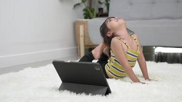 niña asiática acostada en la alfombra con una tableta para jugar gimnasia con postura de arco, use el pie para tocar su cabeza en la alfombra en la sala de estar en casa video