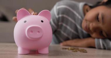 Closeup of Hand girl putting coins into piggy bank while sitting on floor in living room. Front view piggy bank on table. Financial, saving money and investment concept. video