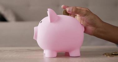 Closeup of Hand girl putting coins into piggy bank while sitting on floor in living room. Side view piggy bank on table. Financial, saving money and investment concept. video