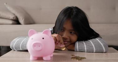 Closeup of Happy Asian girl putting coins into piggy bank while sitting on floor in living room. Smiling young girl saving money. Financial and investment concept. video