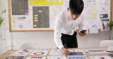 Portrait of Asian man checking to task on workplace desk while standing in office. Male holding smartphone and writing on paper. Man using mobile phone. video