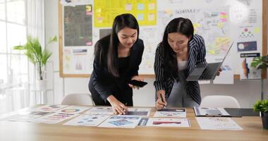 Two asian women checking to task on workplace desk while standing in office. One woman holding laptop and writing on paper, One woman using smartphone and writing notes on colorful sticky papers. video