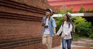 feliz pareja de viajeros asiáticos con la mano del sombrero juntos mientras visitan el antiguo templo. sonriente joven y mujer caminando y mirando templo antiguo. concepto de vacaciones, viajes y pasatiempos. video