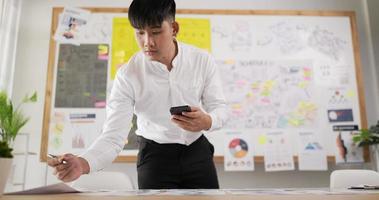 retrato de un hombre asiático revisando la tarea en el escritorio del lugar de trabajo mientras está sentado en la oficina. hombre sosteniendo teléfono inteligente y escribiendo en papel. hombre usando teléfono móvil.